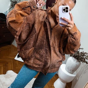 Könnte beinhalten: Braune Lederjacke mit Used-Look, mit Reißverschluss vorne, Taschen und Kragen. Die Jacke hat eine Vintage-Ästhetik und wird mit einer blauen Jeans kombiniert.