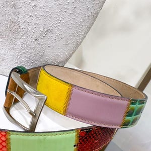 Op de afbeelding: Een patchwork leren riem met een zilverkleurige gesp. De riem heeft delen van geel, roze en groen leer, met een groen reptieltextuur gedeelte. Een stijlvol accessoire, ongeveer 3 cm breed.