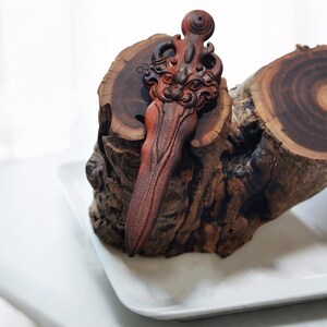May include: A wooden carving of a stylized sword with a face on the hilt. The sword is resting on a piece of wood with a circular grain pattern.