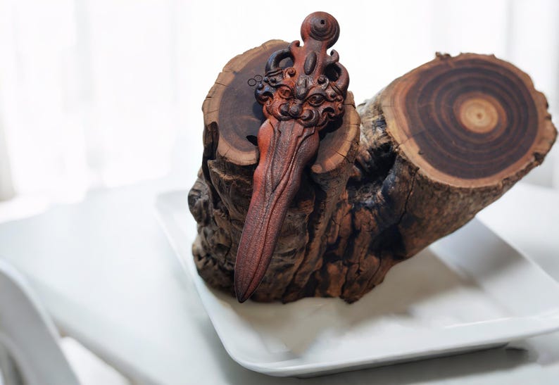 May include: A wooden carving of a stylized sword with a face on the hilt. The sword is resting on a piece of wood with a circular grain pattern.