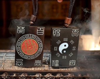 Puede incluir: Dos colgantes rectangulares de madera oscura con diseños detallados. Uno presenta un diseño de moneda roja y dorada, el otro un símbolo de yin-yang blanco. Ambos tienen caracteres chinos y cuelgan de un cordón marrón.