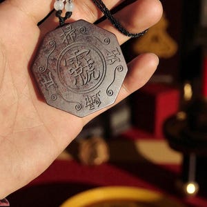 May include: A dark brown, octagonal pendant with Chinese characters carved into its surface. The pendant is suspended from a black cord with white beads. The design features a central circular motif surrounded by ornate details.