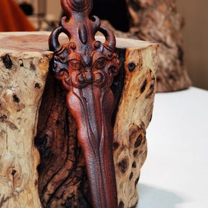 May include: A dark brown wooden carving of a stylized dagger with a face on the handle. The dagger is resting on a piece of wood with a rough, natural texture.