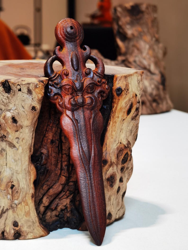 May include: A dark brown wooden carving of a stylized dagger with a face on the handle. The dagger is resting on a piece of wood with a rough, natural texture.
