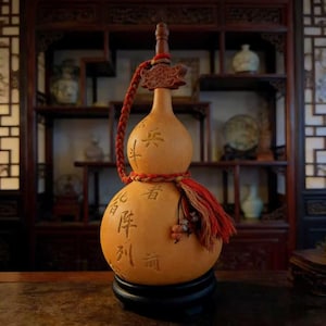 以下が含まれることがあります： 彫刻された中国の文字が施されたひょうたん型の装飾品。木製の栓、赤い三つ編みのコード、タッセルが特徴です。ひょうたんはタン色で、ベースは黒です。背景には、さまざまな装飾品が置かれた木製の棚があります。