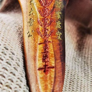 May include: Close-up of a wooden object with intricate carvings and inscriptions in gold and red. The wood has a warm, reddish-brown hue with visible grain. The carvings appear to be of a religious or cultural nature, with detailed patterns and symbols.