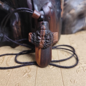 May include: A carved wooden pendant featuring a detailed face with a beard and flowing hair. The pendant is suspended from a dark brown cord. The wood has a natural grain and a mix of light and dark brown tones.