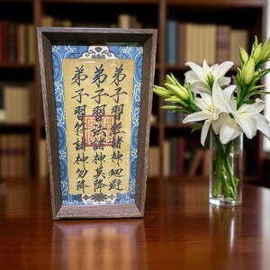 May include: A framed artwork with Chinese calligraphy in a wooden frame, displayed on a dark wooden table. A vase of white lilies sits to the right. The background features a bookshelf filled with books.