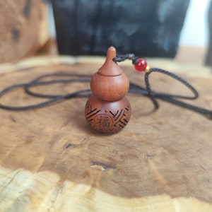 May include: A brown wooden gourd pendant necklace. The pendant has a carved design and the Chinese character for "blessing." It hangs on a black cord with a red bead. The necklace is displayed on a wooden surface.