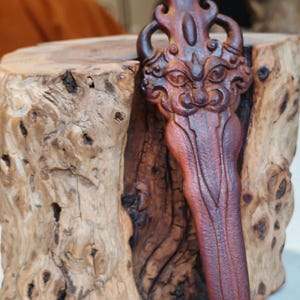 May include: A hand-carved wooden dagger with a detailed face and intricate design. The dagger is a dark reddish-brown color and is resting on a wooden stump.