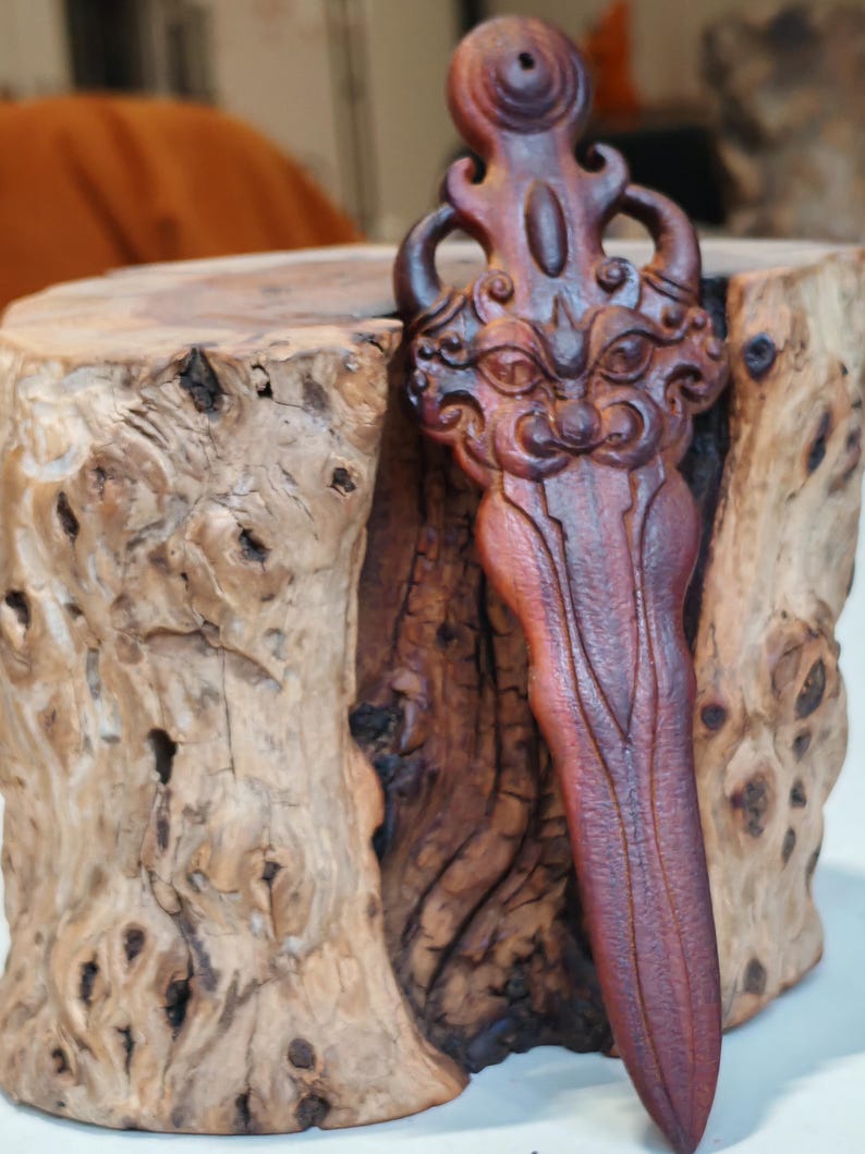 May include: A hand-carved wooden dagger with a detailed face and intricate design. The dagger is a dark reddish-brown color and is resting on a wooden stump.