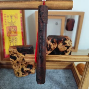 May include: A dark brown, cylindrical wooden object with a red cord hanging from the top. The object is suspended in front of a wooden frame and other wooden decorative pieces. The wood has a textured, natural appearance.