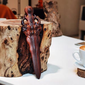 May include: A wooden sculpture of a stylized sword with a face carved into the blade. The sculpture is resting on a wooden stump.