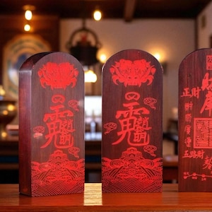 May include: Three wooden plaques with red Chinese characters and decorative patterns. The plaques are rectangular with rounded tops and stand upright on a wooden surface. The background is blurred, suggesting an interior setting.