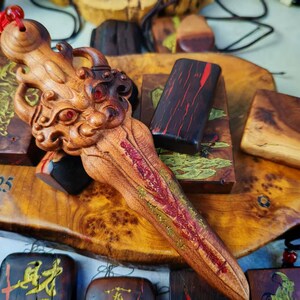 May include: A detailed wooden carving of a dragon-like creature with red accents and a red tassel. The carving is lying on a wooden surface with other wooden carvings and a black cord. The text "a 2025" is visible.