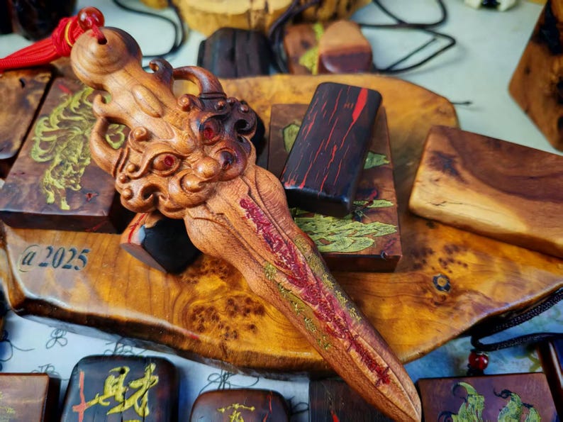 May include: A detailed wooden carving of a dragon-like creature with red accents and a red tassel. The carving is lying on a wooden surface with other wooden carvings and a black cord. The text "a 2025" is visible.