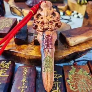 May include: A carved wooden pendant with a detailed face design at the top and Chinese characters. The pendant is a light brown color with red and gold accents. A red cord is attached to the pendant. Other wooden carvings are in the background.