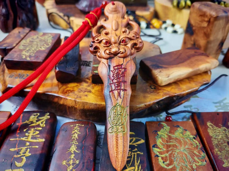 May include: A carved wooden pendant with a detailed face design at the top and Chinese characters. The pendant is a light brown color with red and gold accents. A red cord is attached to the pendant. Other wooden carvings are in the background.