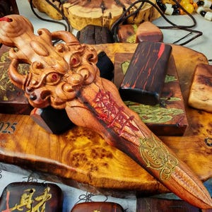 May include: A carved wooden dagger with a dragon head handle and intricate designs. The dagger is light brown with red and gold accents. Other wooden items are in the background, including a red tassel and a wooden box with the text "@2025".