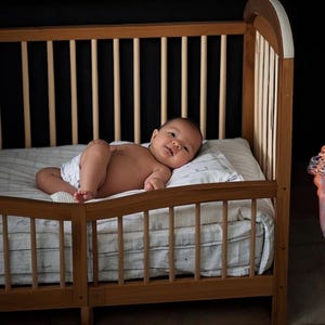 May include: A baby lying in a wooden crib with white bedding. The baby is wearing a diaper and has a relaxed expression.