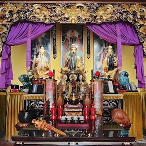 May include: A detailed display featuring multiple golden statues, ornate carvings, and vibrant purple drapes. The scene includes a central altar with various religious artifacts, set against a backdrop of framed portraits and a golden decorative arch.