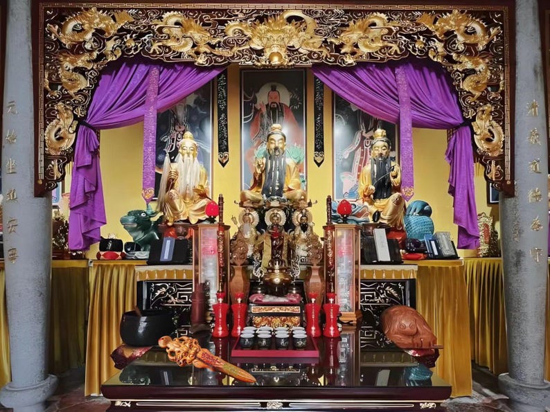 May include: A detailed display featuring multiple golden statues, ornate carvings, and vibrant purple drapes. The scene includes a central altar with various religious artifacts, set against a backdrop of framed portraits and a golden decorative arch.