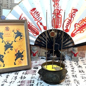 Puede incluir: Una obra de arte enmarcada con figuras negras sobre un fondo amarillo. Varillas de incienso en un incensario de bronce, con un abanico decorativo detrás. Dos estatuas de leones de piedra flanquean el incensario, y se ve caligrafía.