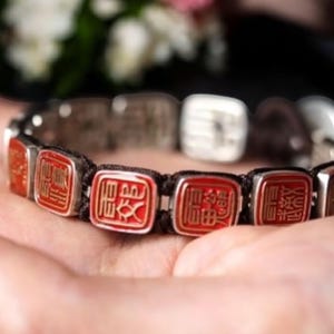 Könnte beinhalten: Ein silbernes Armband mit roten rechteckigen Anhängern. Jeder Anhänger zeigt ein goldfarbenes Symbol. Das Armband ist an einem schwarzen Band befestigt. Der Hintergrund ist verschwommen, mit Andeutungen von weißen Blumen.