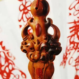 May include: A carved wooden pendant with a detailed design. The pendant features a dragon-like figure with intricate carvings and a red bead at the top. The wood has a warm, reddish-brown hue, and the background is a blurred white with red accents.
