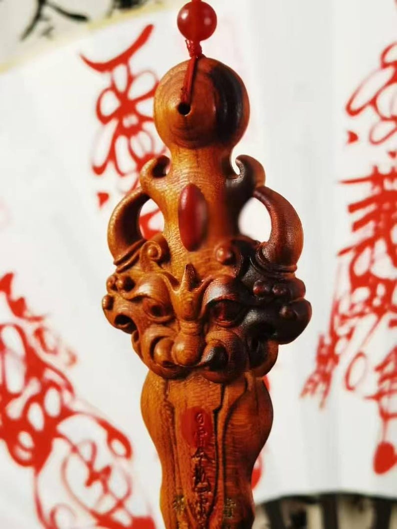 May include: A carved wooden pendant with a detailed design. The pendant features a dragon-like figure with intricate carvings and a red bead at the top. The wood has a warm, reddish-brown hue, and the background is a blurred white with red accents.