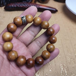 Könnte beinhalten: Ein Holzperlenarmband mit einem silbernen Akzent mit einem Yin-Yang-Symbol. Die Perlen sind warmbraun und aufgefädelt. Das Armband wird in einer Hand gehalten.