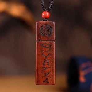 Puede incluir: Colgante rectangular de madera marrón rojizo con diseños grabados, suspendido de un cordón negro. El colgante presenta un diseño circular tallado en la parte superior y patrones lineales a lo largo del cuerpo. Una cuenta roja adorna el cordón.