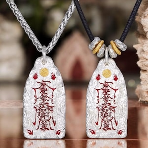 Puede incluir: Dos colgantes de plata con cadenas negras y plateadas. Los colgantes son rectangulares y presentan un fondo plateado con caracteres chinos rojos y dorados. Los colgantes están sobre una superficie de madera.