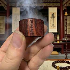 Puede incluir: Un pequeño quemador de incienso cilíndrico de madera con caracteres chinos tallados. La madera marrón oscuro tiene vetas visibles. El humo blanco se eleva desde la parte superior, lo que indica su uso. El quemador se sostiene en una mano.