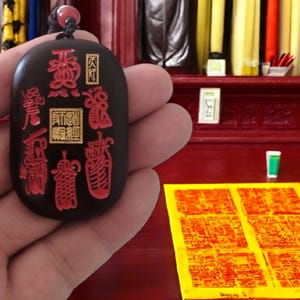 Könnte beinhalten: Ein schwarzer Jade-Anhänger mit roten und goldenen chinesischen Schriftzeichen. Der Anhänger wird in einer Hand gehalten und befindet sich vor einer roten Holzoberfläche.