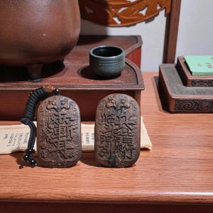 May include: Two brown, carved wooden pendants with intricate Chinese characters. One pendant has a black cord with beads. The pendants rest on a wooden table, with a small ceramic cup and other decorative items in the background.