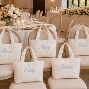 May include: Five off-white quilted tote bags with two handles each, featuring embroidered names in light blue script. The names Rose, Mary, Sarah, Emily, and Olivia are stitched onto the front of each individual bag, which are displayed on chairs.