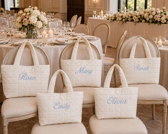 May include: Five off-white quilted tote bags with two handles each, featuring embroidered names in light blue script. The names Rose, Mary, Sarah, Emily, and Olivia are stitched onto the front of each individual bag, which are displayed on chairs.