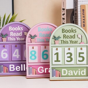 May include: Three wooden book trackers in purple, pink, and green, each with a curved top and the words "Books Read This Year." Each tracker displays a different number of books read, with names below. A small potted plant decorates the top.