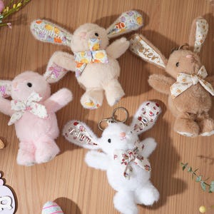 May include: Four plush bunny toys in pink, white, and tan, with floral patterned bows and ears. Each bunny has a name embroidered on its ear. One white bunny is a keychain. Easter-themed.