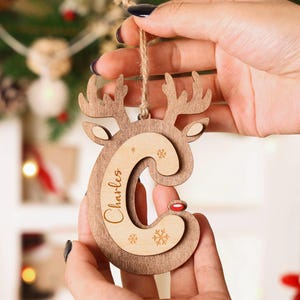 May include: Wooden Christmas ornament in the shape of a reindeer with the letter "C" cut out. The name "Charles" is engraved on the ornament. The reindeer has antlers, ears, and a red nose. The ornament is held by a string.