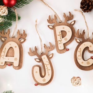 Può includere: Ornamenti natalizi in legno a forma di renna, ognuno con una lettera e un nome: Andrea, Phoebe, Charles e Joshua. Gli ornamenti hanno un centro in legno chiaro e un contorno in legno più scuro, con dettagli di fiocchi di neve e nasi rossi. Sono appesi a uno spago.