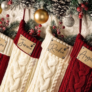 May include: Four personalized Christmas stockings in red and cream cable knit with names Claire, Jerome, Richie, and Angela on leather tags. Festive decor includes ornaments, pine boughs, and berries.