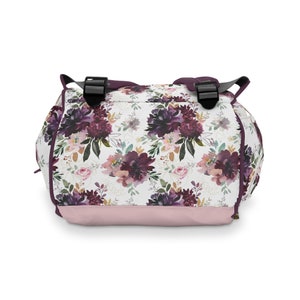 May include: A pink and white floral backpack with purple flowers and gold accents. The backpack has two adjustable straps and a zipper closure.