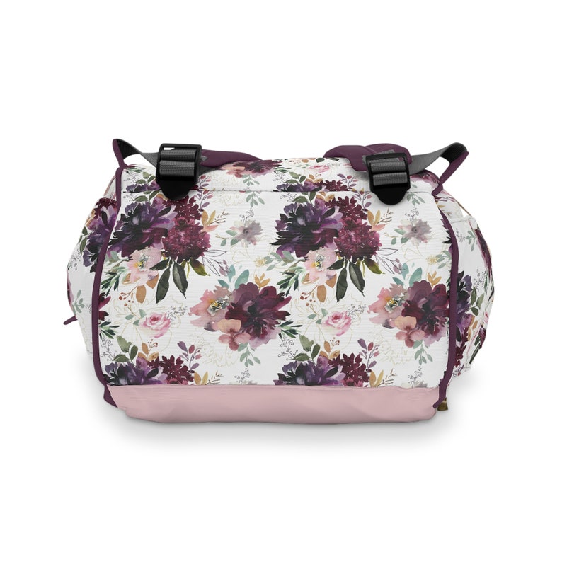 May include: A pink and white floral backpack with purple flowers and gold accents. The backpack has two adjustable straps and a zipper closure.