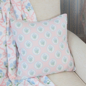 May include: A pink square pillow with a repeating pattern of blue floral medallions. The pillow is on a cream-colored chair, with a matching blanket featuring blue flowers and pink ribbons. The overall theme is floral and feminine.