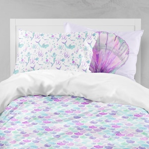 May include: A white duvet cover with a mermaid scale pattern in shades of purple, blue, and green. Two pillowcases are shown, one with a mermaid and sea creature design and the other with a large seashell.