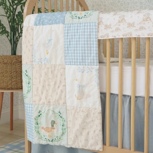 May include: A blue and cream patchwork baby quilt with a duck design. The quilt is draped over a wooden cot with a blue and white gingham cot skirt.