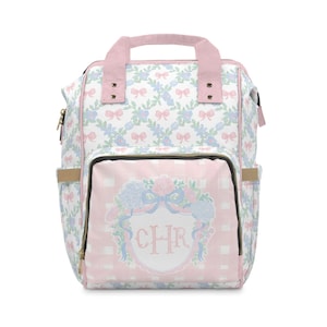 May include: A white and pink floral print backpack with a pink handle and straps. The front pocket features a pink and white gingham pattern with a floral crest and the initials "CHR". The backpack has gold zippers and side pockets.