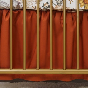 Op de afbeelding: Een roestkleurige wieg rok met een gerimpelde rand hangt aan een gouden metalen wieg frame.